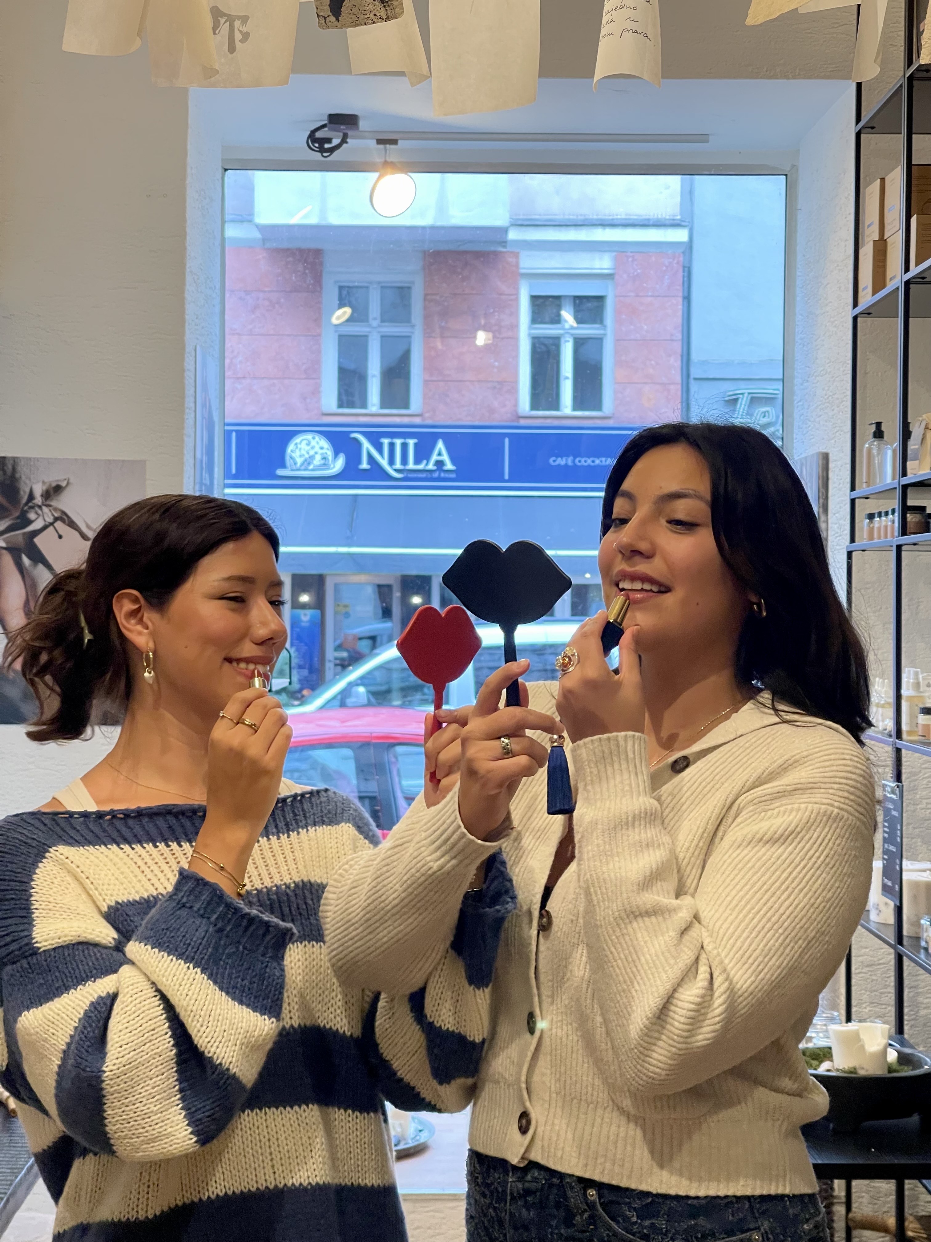 Two women apply lipstick using mirrors.