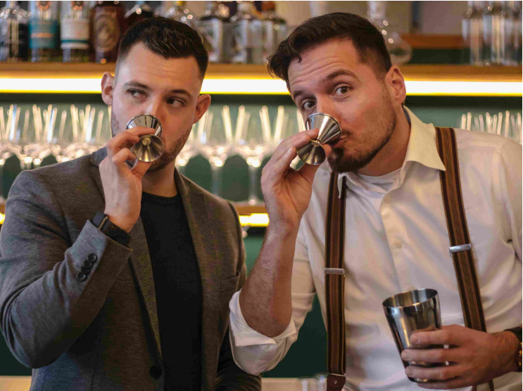 Two men smell metal jiggers in bar.