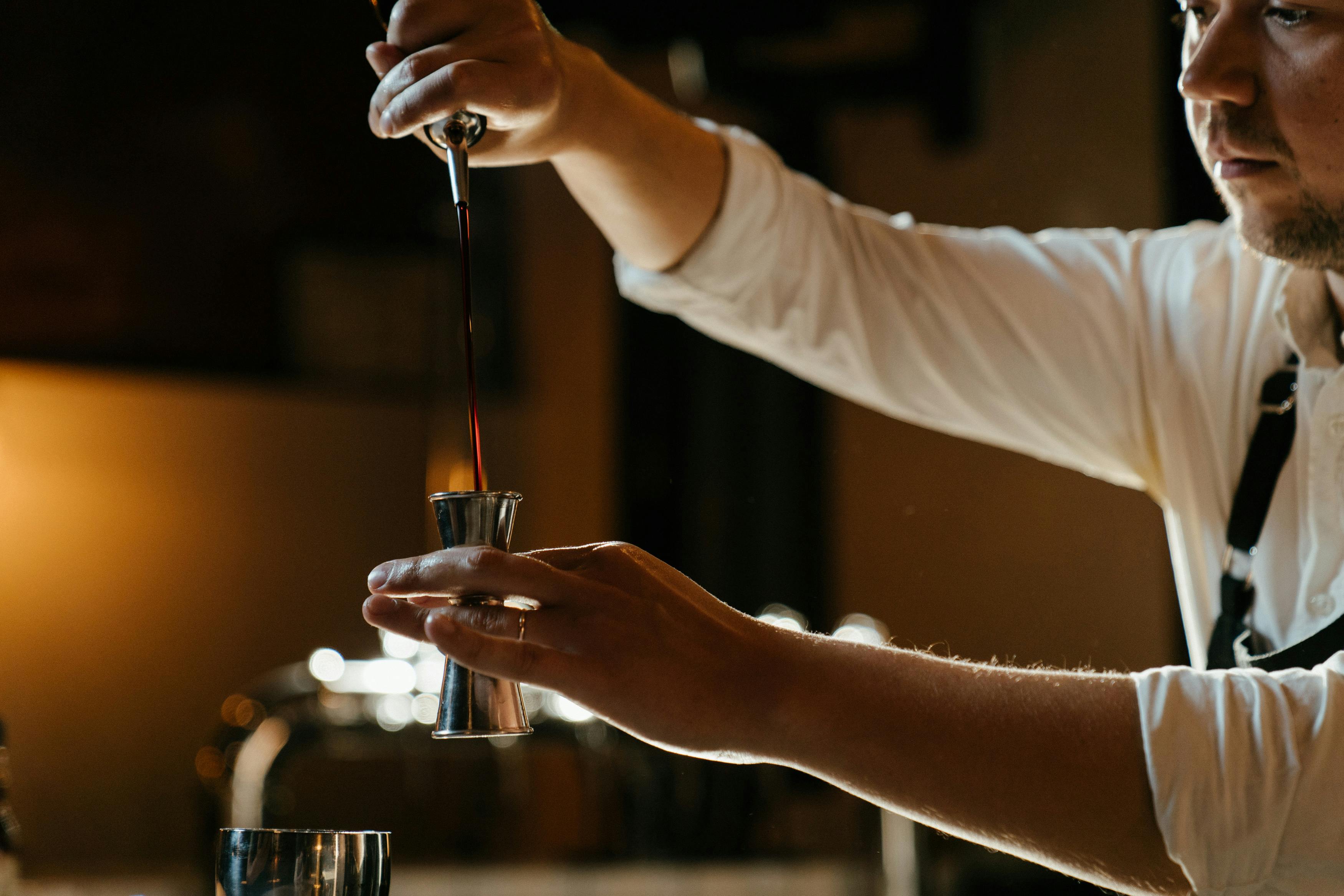 Barkeeper gießt Flüssigkeit in Jigger, schummrige Barbeleuchtung.