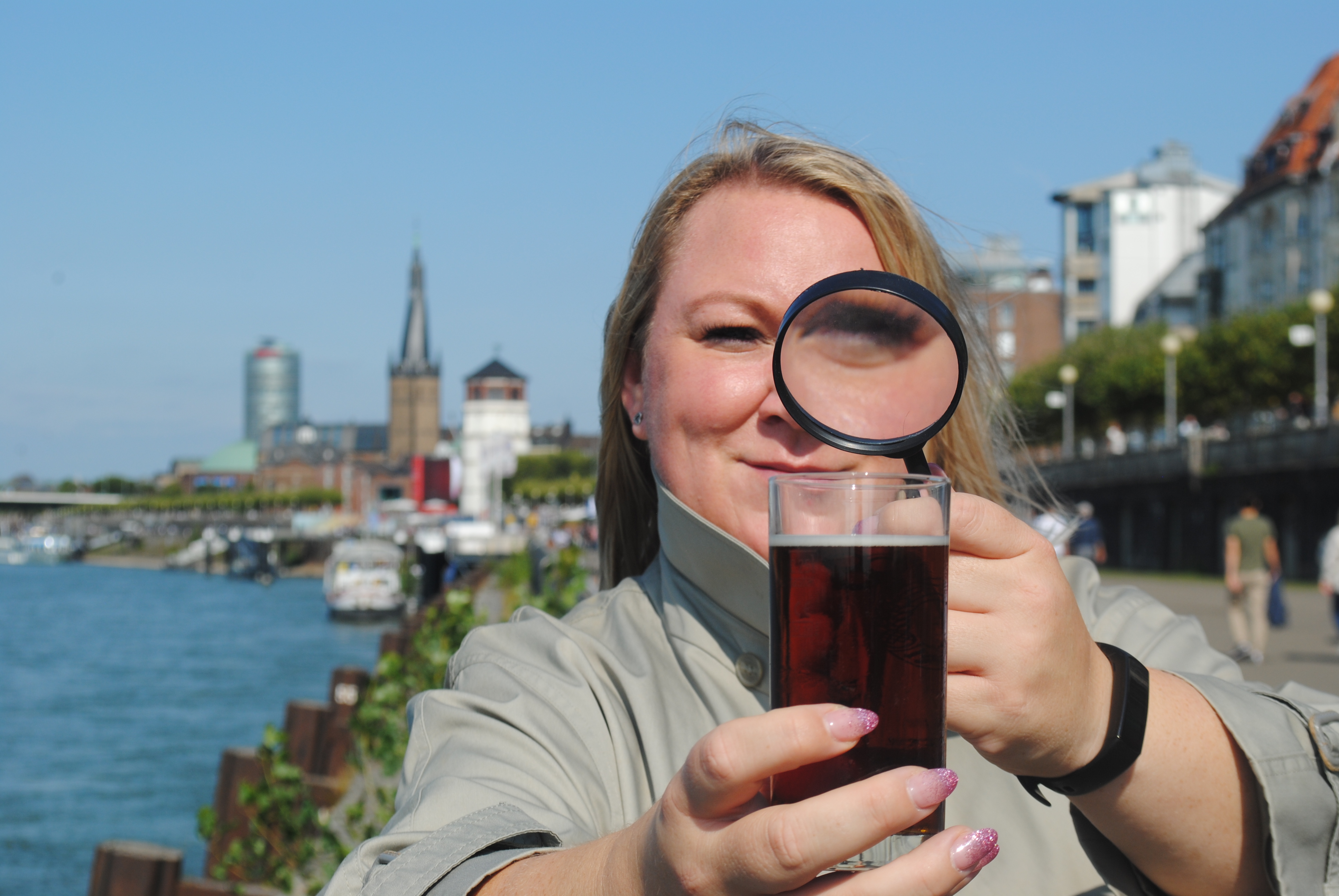 Woman holds magnifying glass over drink by river.