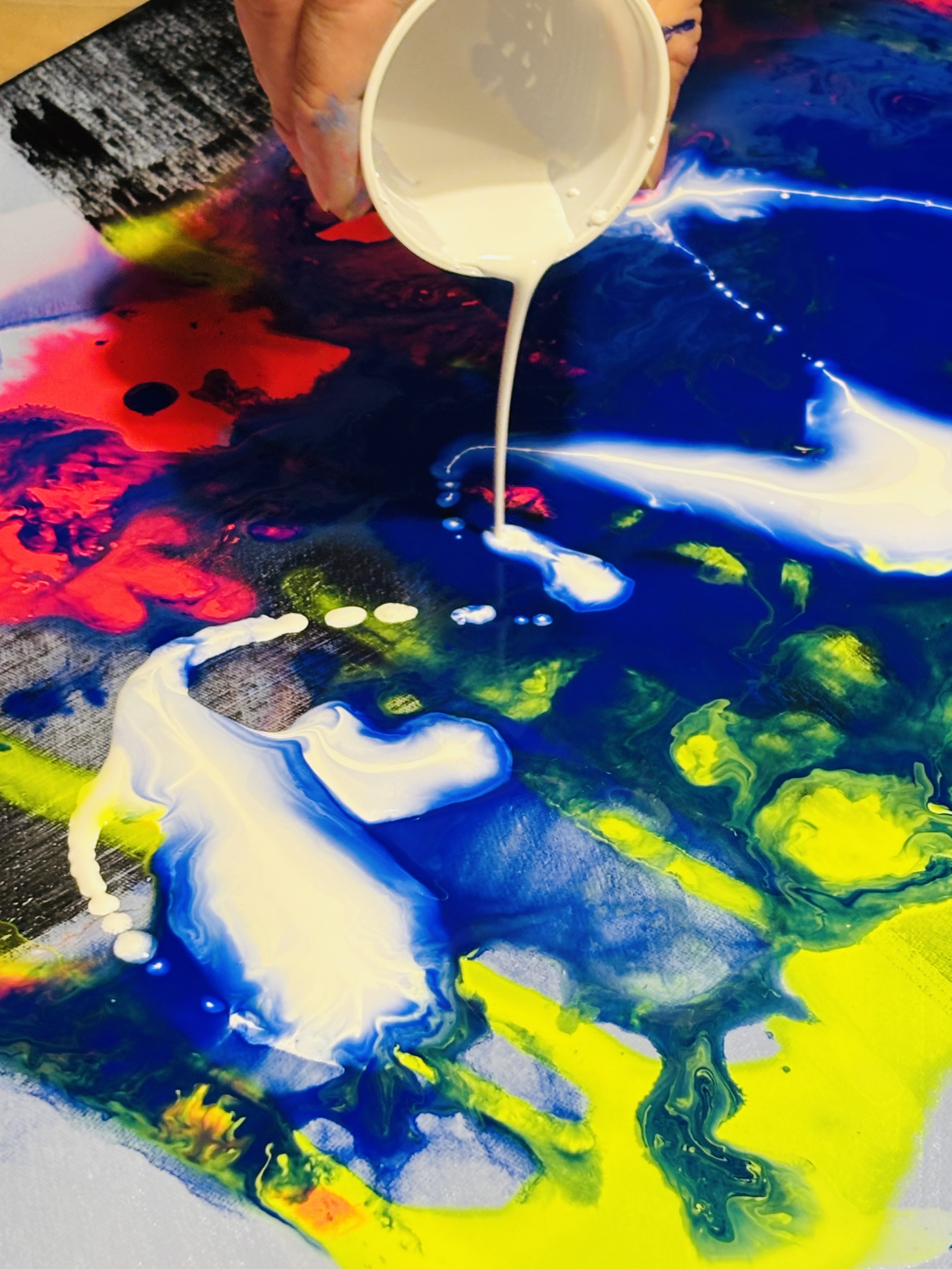 Hand pours white paint on colorful canvas.