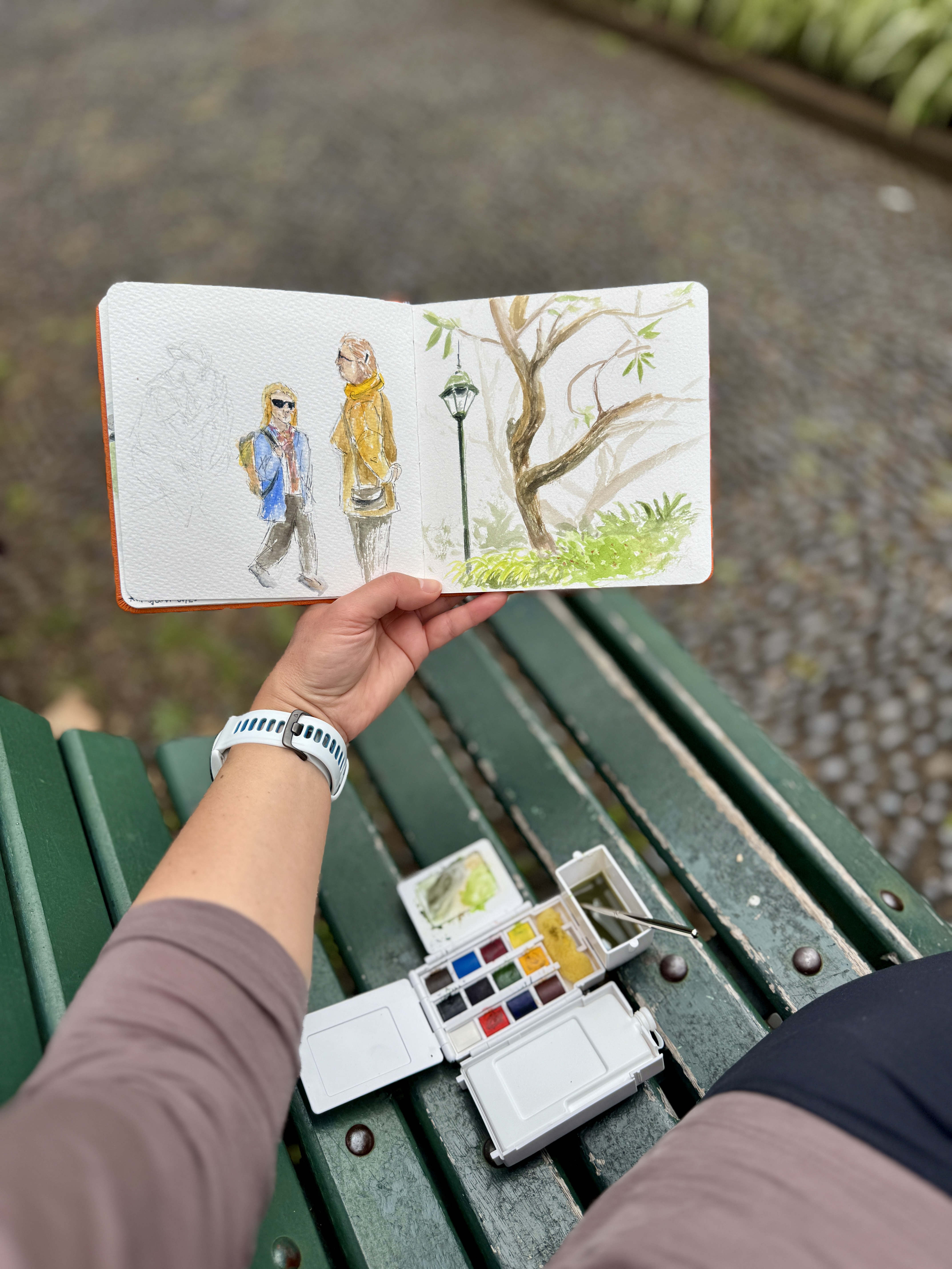 Hand holding sketchbook with watercolor painting outdoors.