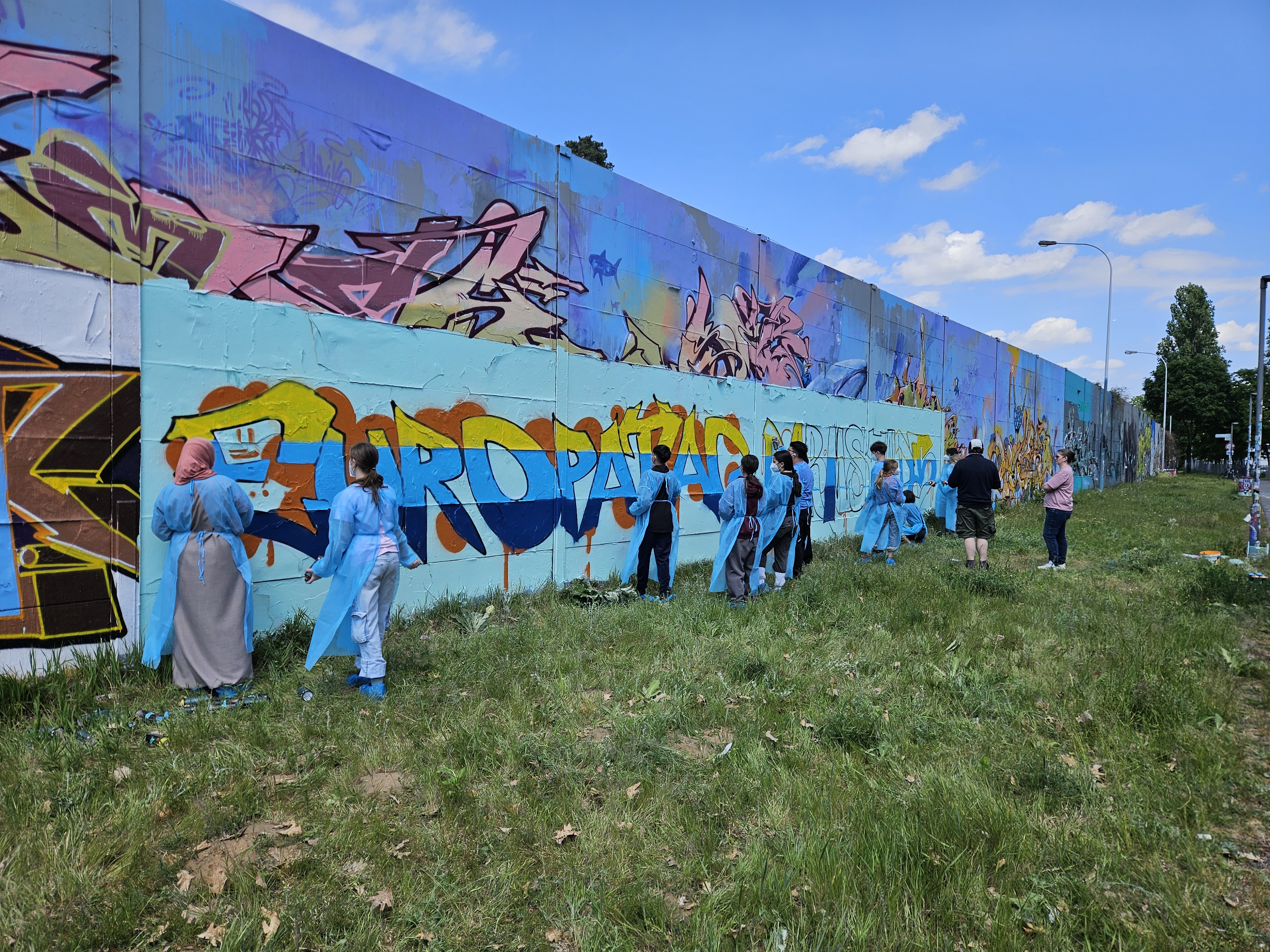 People painting graffiti on a long wall outside.