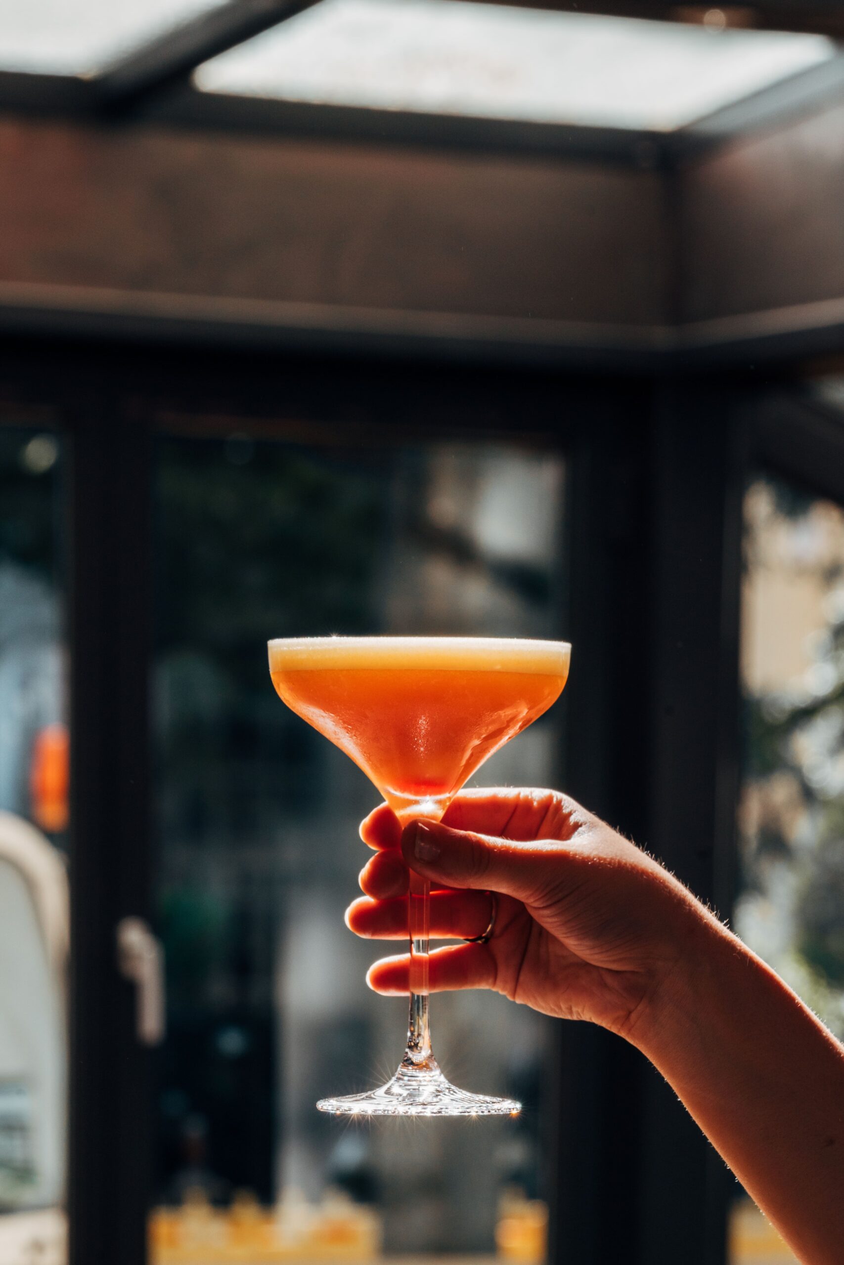 Ein Cocktail wird in der Hand gehalten, vor Fenster.