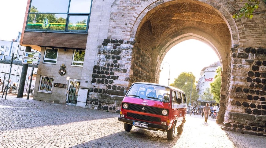 Roter VW-Bus fährt durch historischen Torbogen in Stadt.