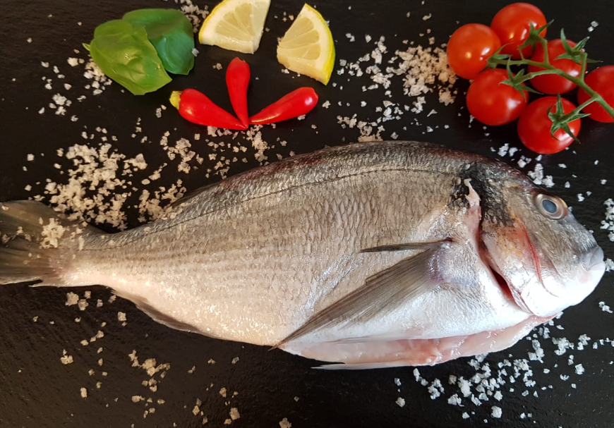A fish is lying on a black stone surrounded by ingredients.