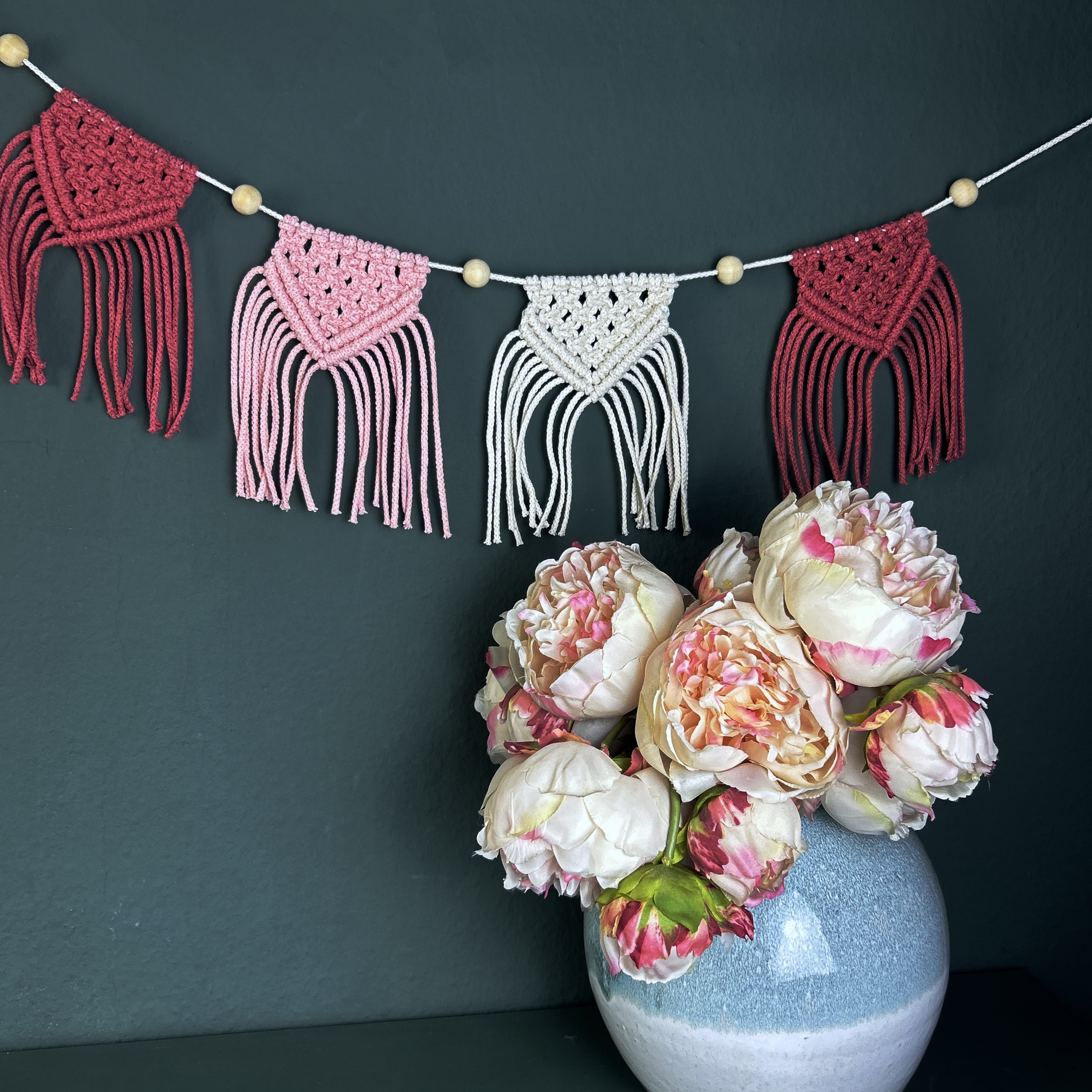 Bunte Makramee-Girlande hängt über einer Blumenarrangierung.