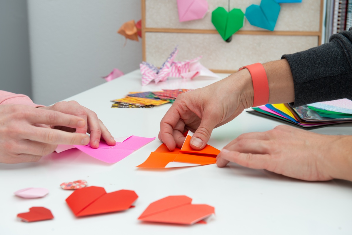 Two hands fold origami hearts made of colorful paper at the table.