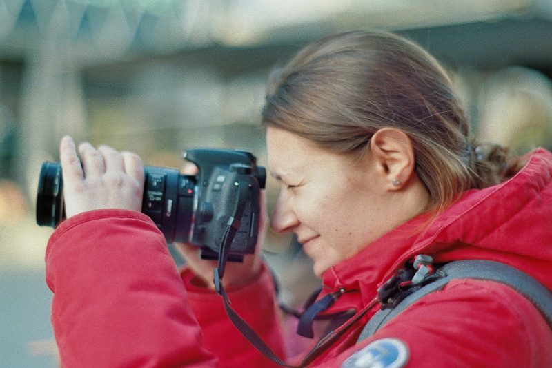 Frau in rotem Anorak fotografiert mit Kamera in urbaner Umgebung.
