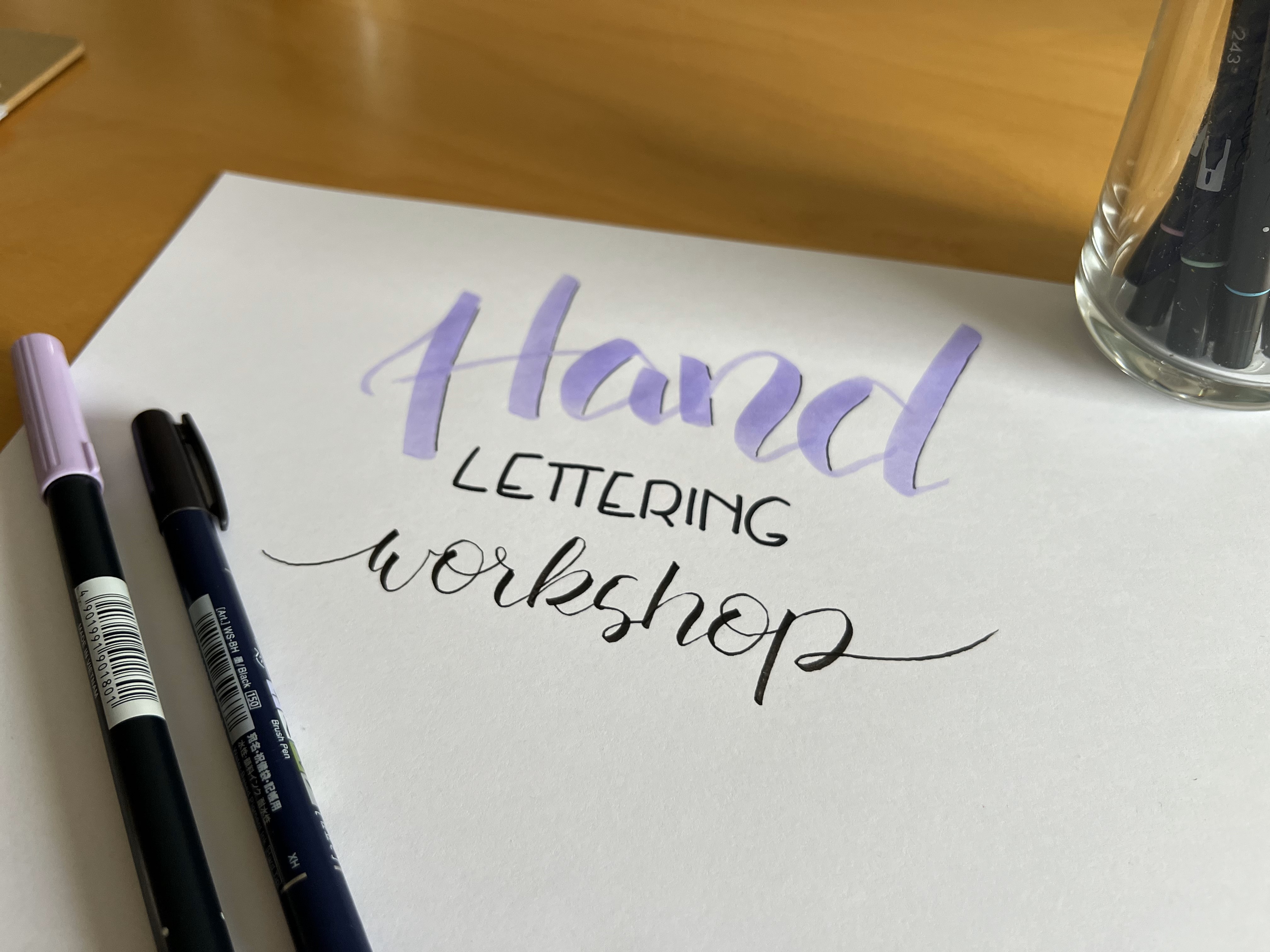 Papier mit `Hand Lettering Workshop`, Stifte daneben, Holzuntergrund.