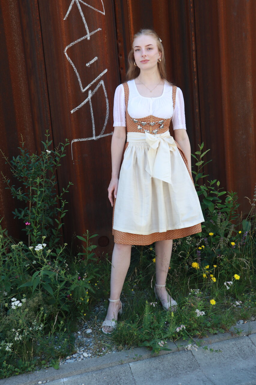 Woman wearing a dirndl stands in front of a rusty wall surrounded by flowers.