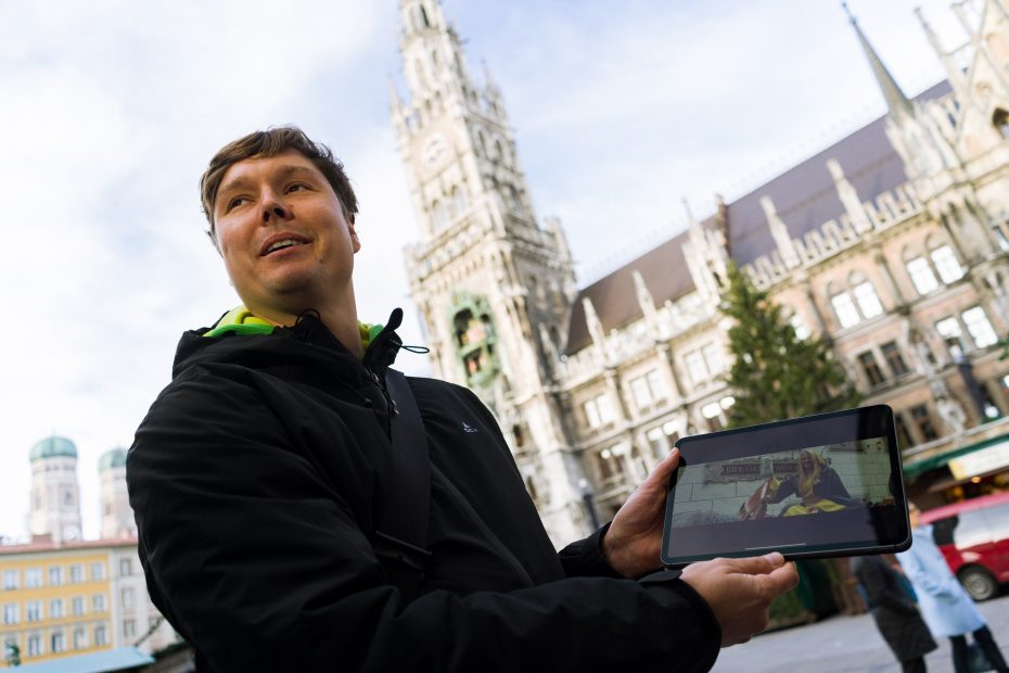 Mann hält Tablet mit Bild vor historischem Gebäude.