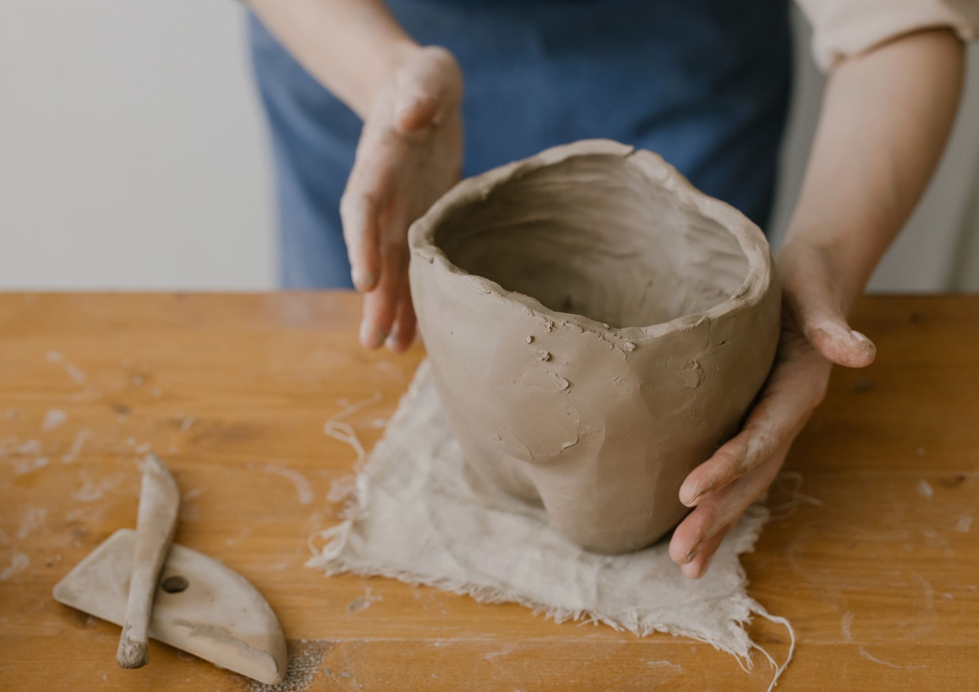 Hands shaping clay pot on wooden table.