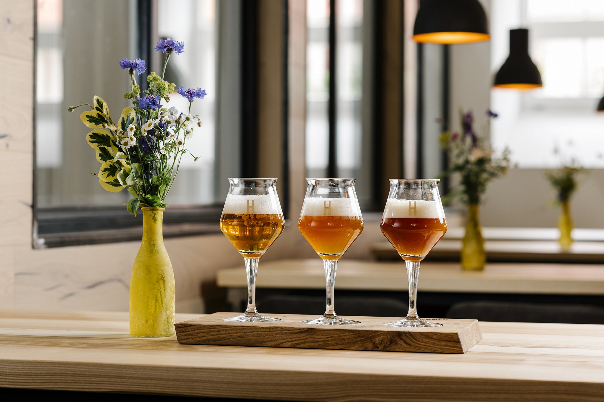 Three beer glasses on wooden tray, floral vase nearby.