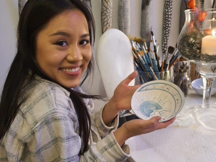 Woman smiling, holding painted plate, near art supplies.
