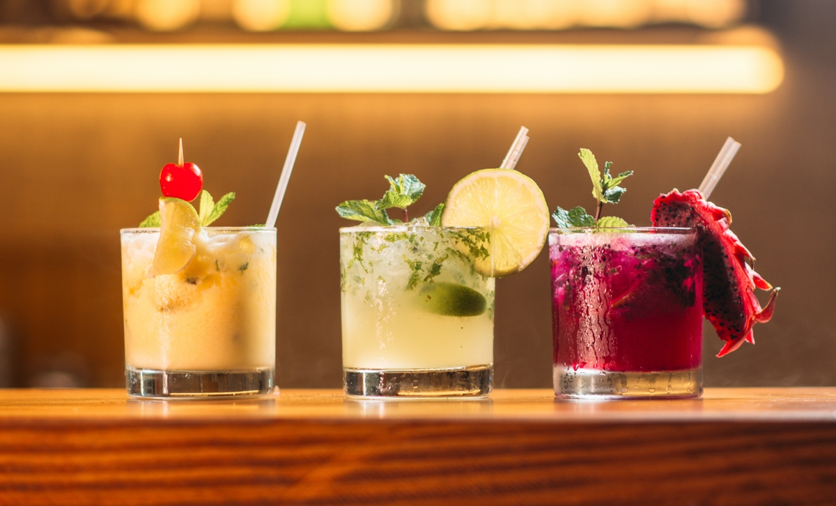 Drei bunte Cocktails stehen auf einem Holzstamm.
