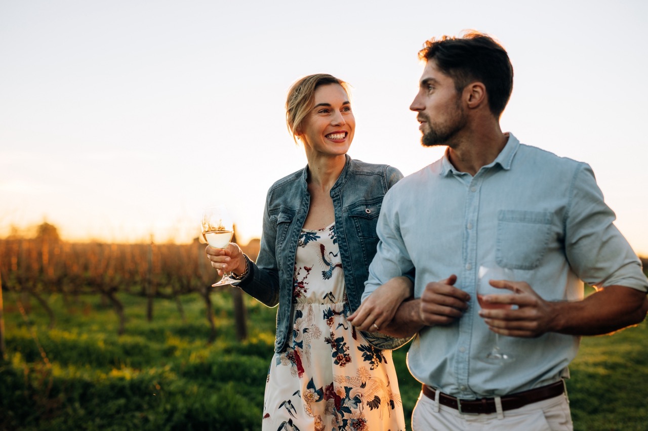 Couple walking in vineyard at sunset, holding wine.