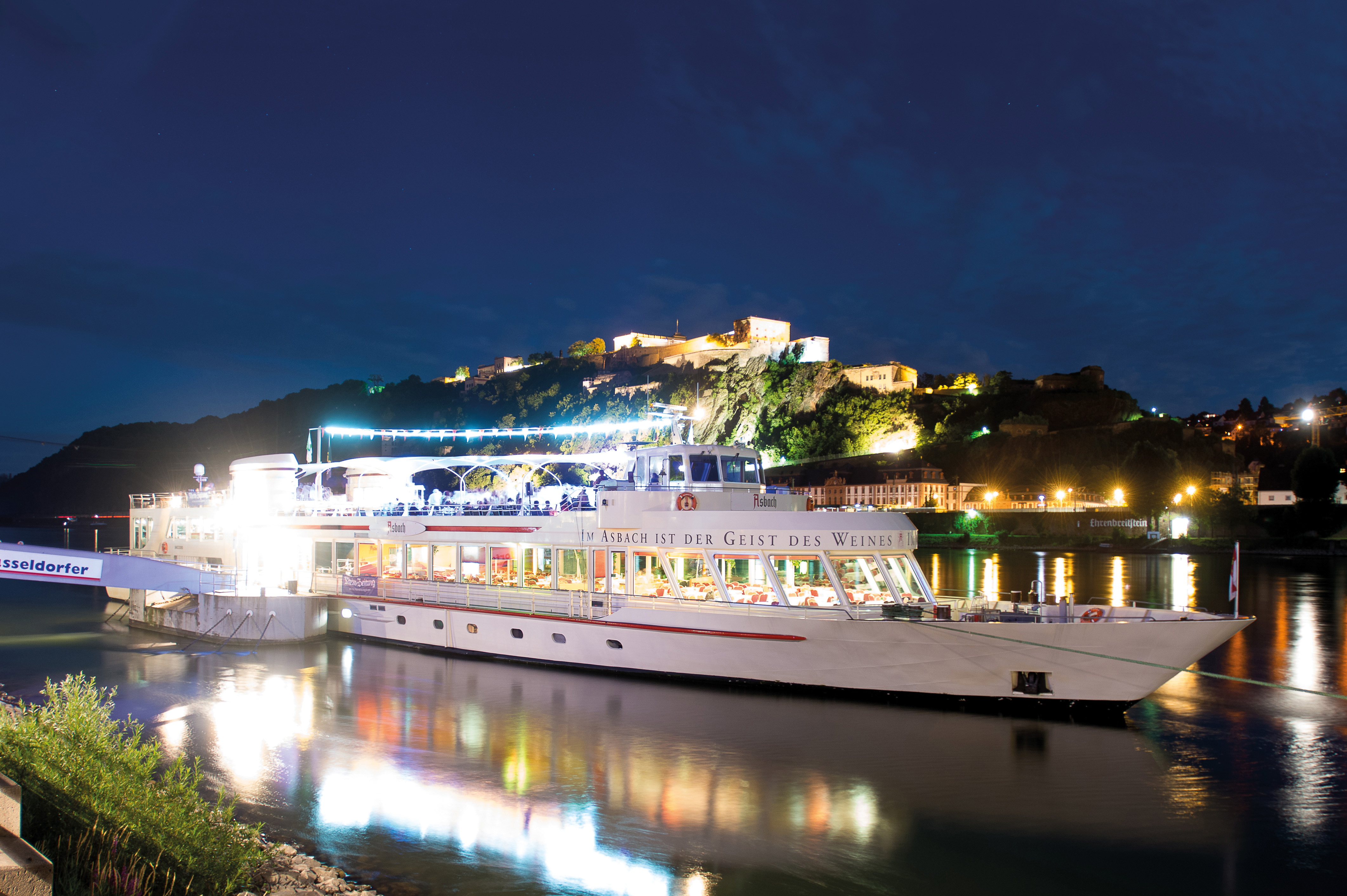 Illuminated boat on river with hills behind.