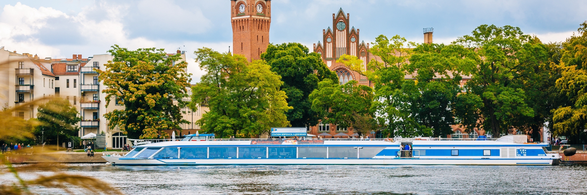 Touristenboot fährt nahe historische Gebäuden, Bäume.