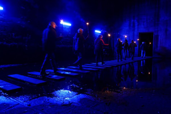 People walk on wooden planks across a dark, reflective waterway.